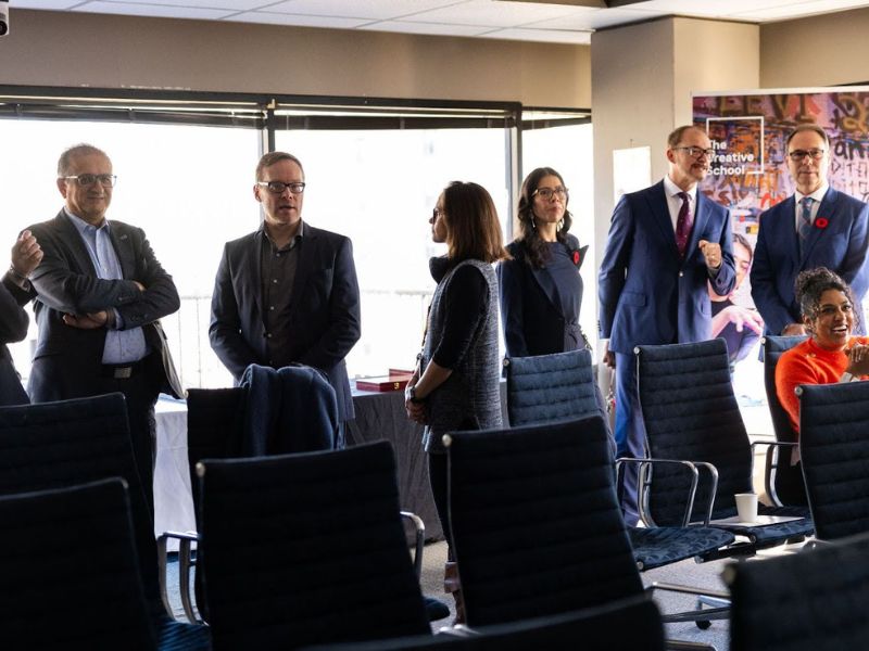 A group of seven people standing and conversing in a conference room, with empty chairs in the foreground. One individual is sitting and smiling.